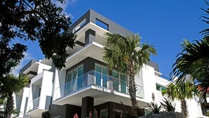 Een modern meerhoog appartementencomplex met balkons, omringd door palmbomen en andere begroeiing.