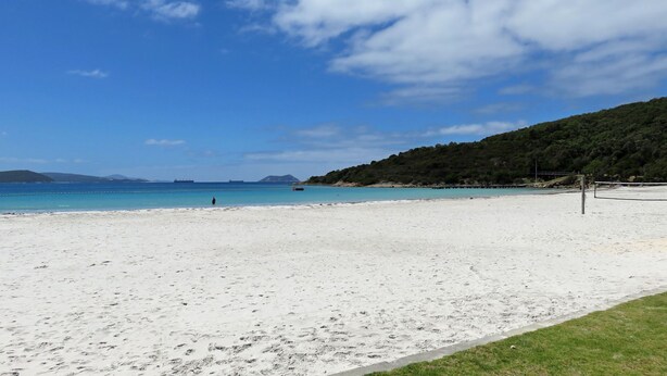 Ein Strand mit weißem Sand, klarem, blauem Wasser und einer hügeligen Landschaft im Hintergrund.
