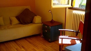 A room with a wooden floor, a sofa with cushions, a small table with a lamp, and a window with curtains. - SUNNY HOUSE, LAND RAISED (Aubenas)