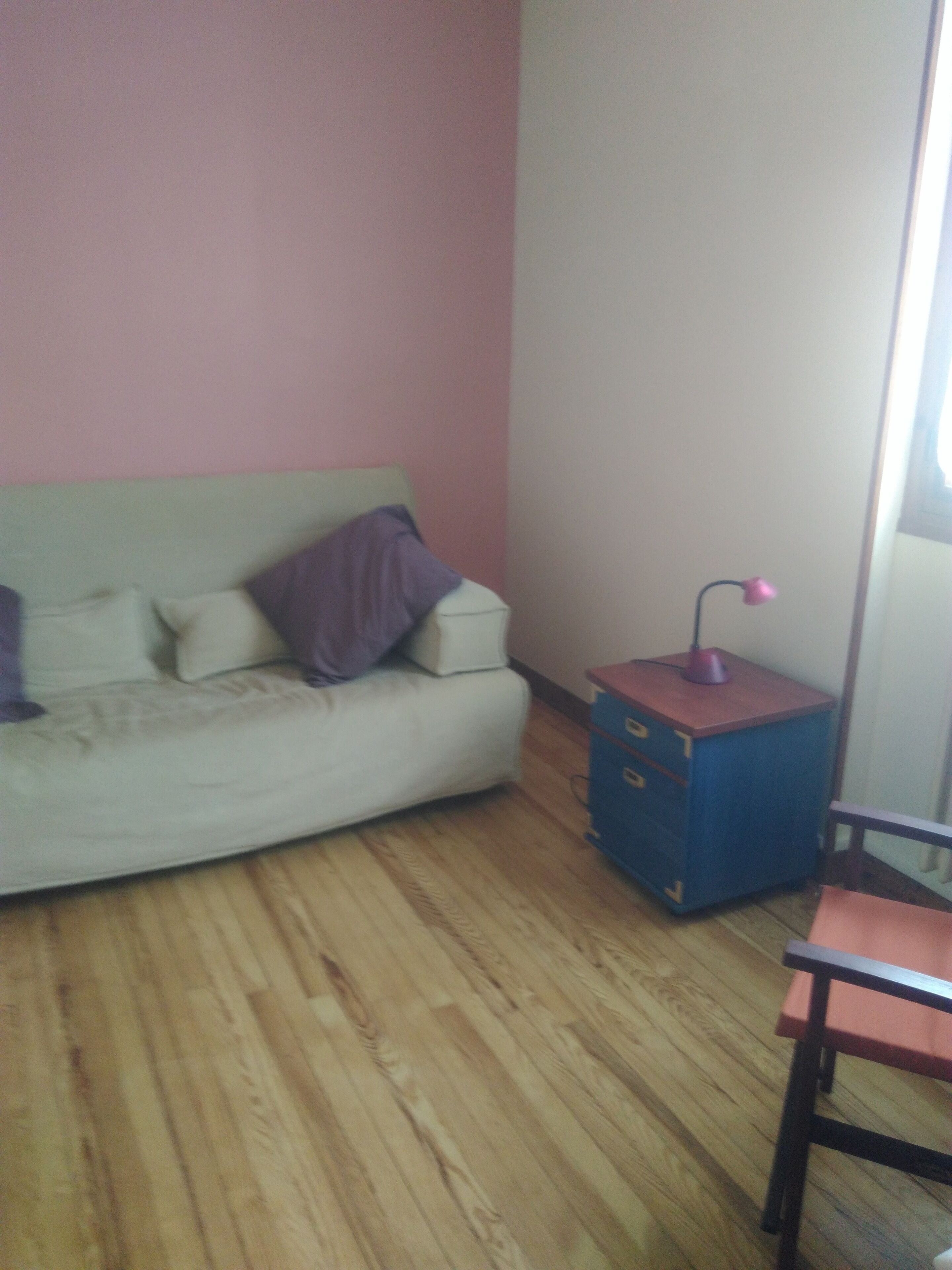 Een kamer met een lichtkleurige bank tegen een roze muur, een klein blauw kastje met lades en een houten vloer.