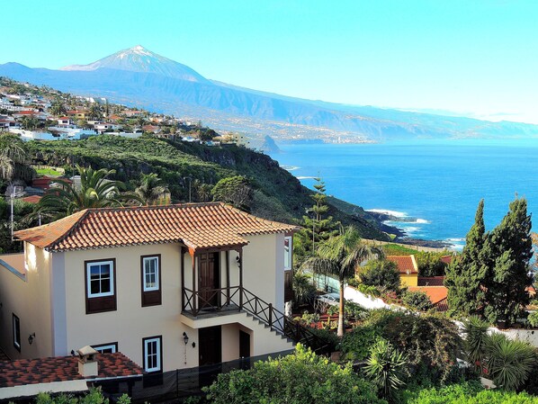 A coastal building with a red-tiled roof, a balcony, and a view of the ocean and a mountain in the background.