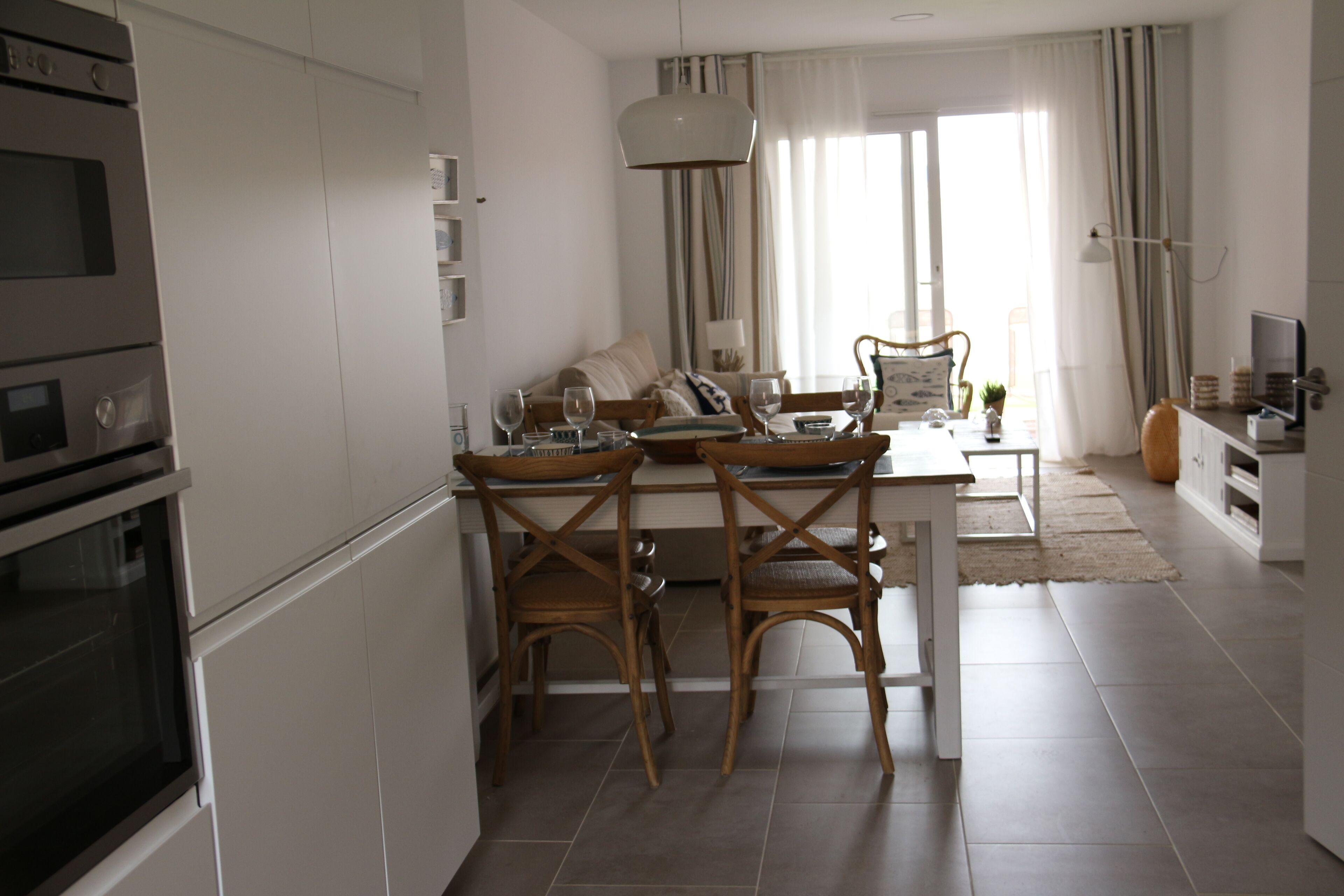 Een moderne keuken met witte kastjes, een eethoek met houten stoelen en een groot raam met gordijnen.