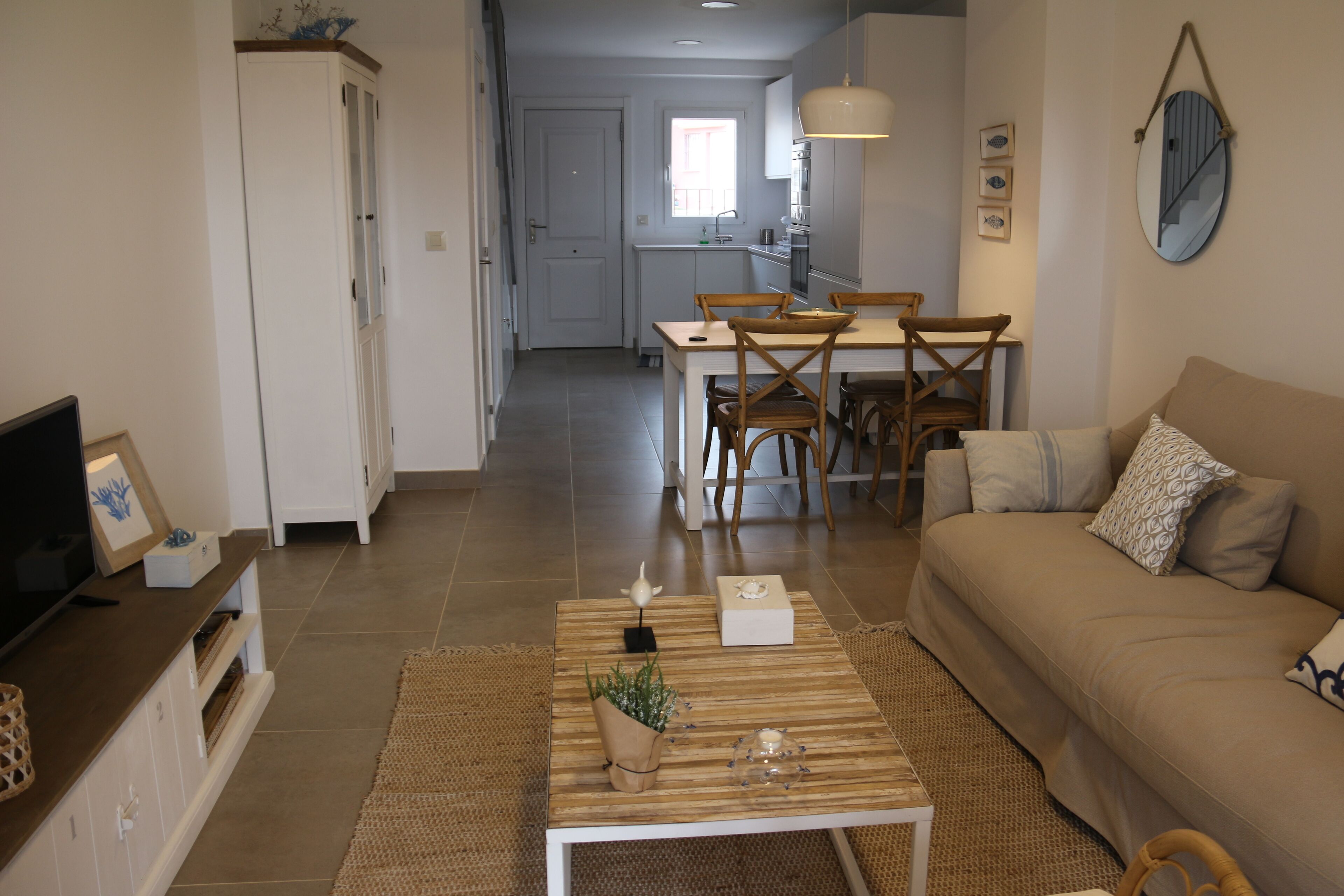 Een moderne woonkamer met een bank, salontafel en tv-meubel. Er is een zithoek met een tafel en stoelen. De keuken is op de achtergrond zichtbaar.