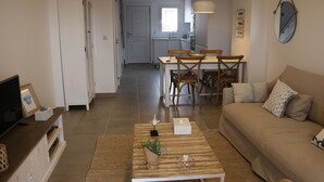 Una sala moderna con sofá, mesa de centro y mueble para televisor. Se observa una zona de comedor con mesa y sillas. La cocina se ve al fondo.