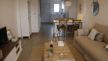 Een moderne woonkamer met een bank, salontafel en tv-meubel. Er is een zithoek met een tafel en stoelen. De keuken is op de achtergrond zichtbaar.