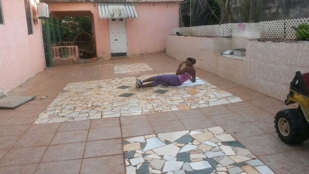 A person lying on a tiled floor in a courtyard with a pink wall, a door, and a small vehicle in the background. - Large home for retreat and family vacation (Mandeville)
