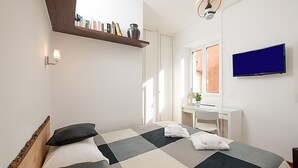 Un dormitorio con una cama, un escritorio, un televisor y una estantería con libros y un florero.