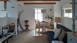 A spacious living room with a dark-colored sofa, a dining table with chairs, a lamp, and a large window providing natural light. - Casa da Bica D'Água - Arelho (Arelho)