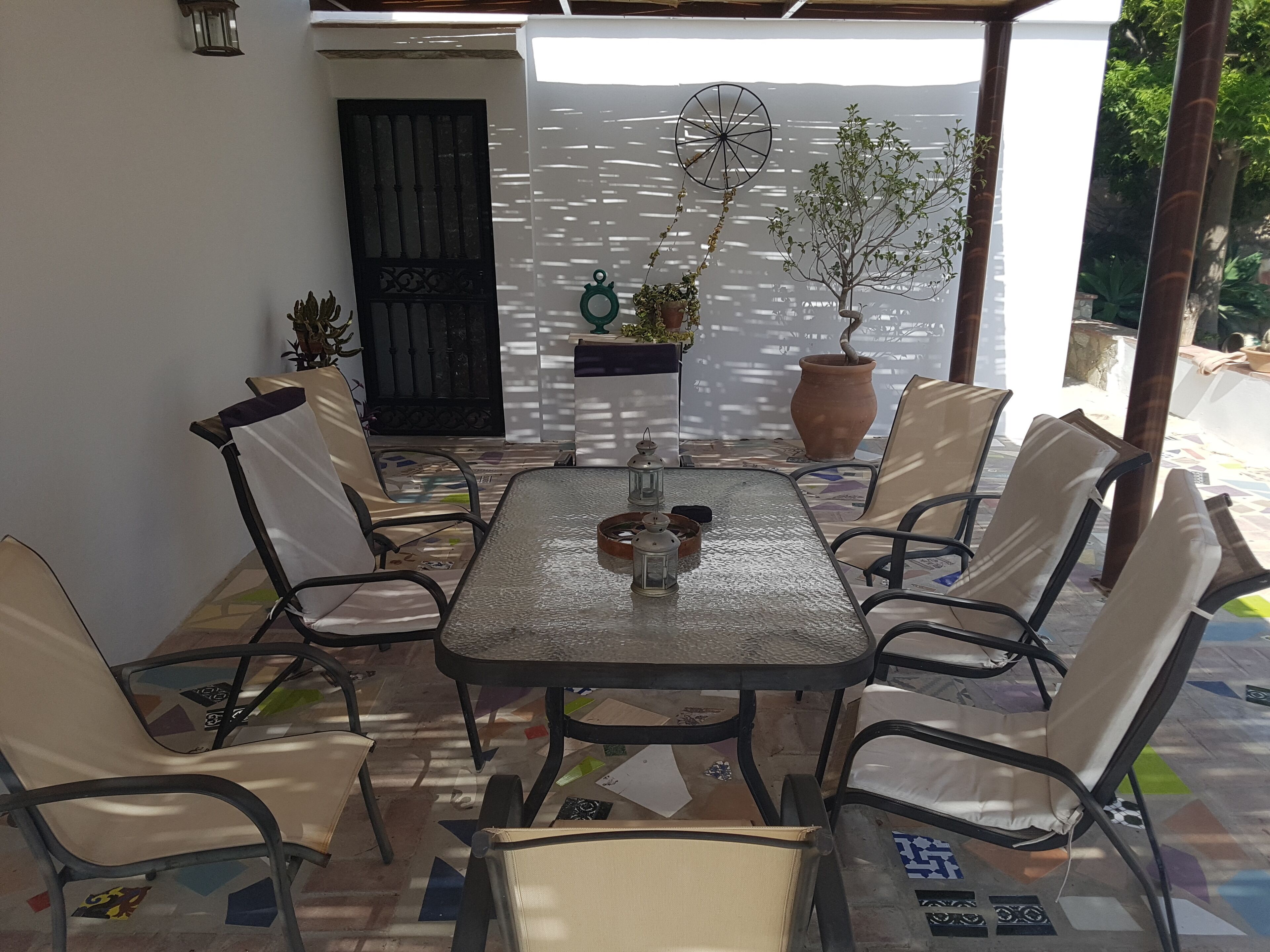Eine überdachte Terrasse mit Tisch und Stühlen, umgeben von Topfpflanzen und einer dekorativen Windglocke.