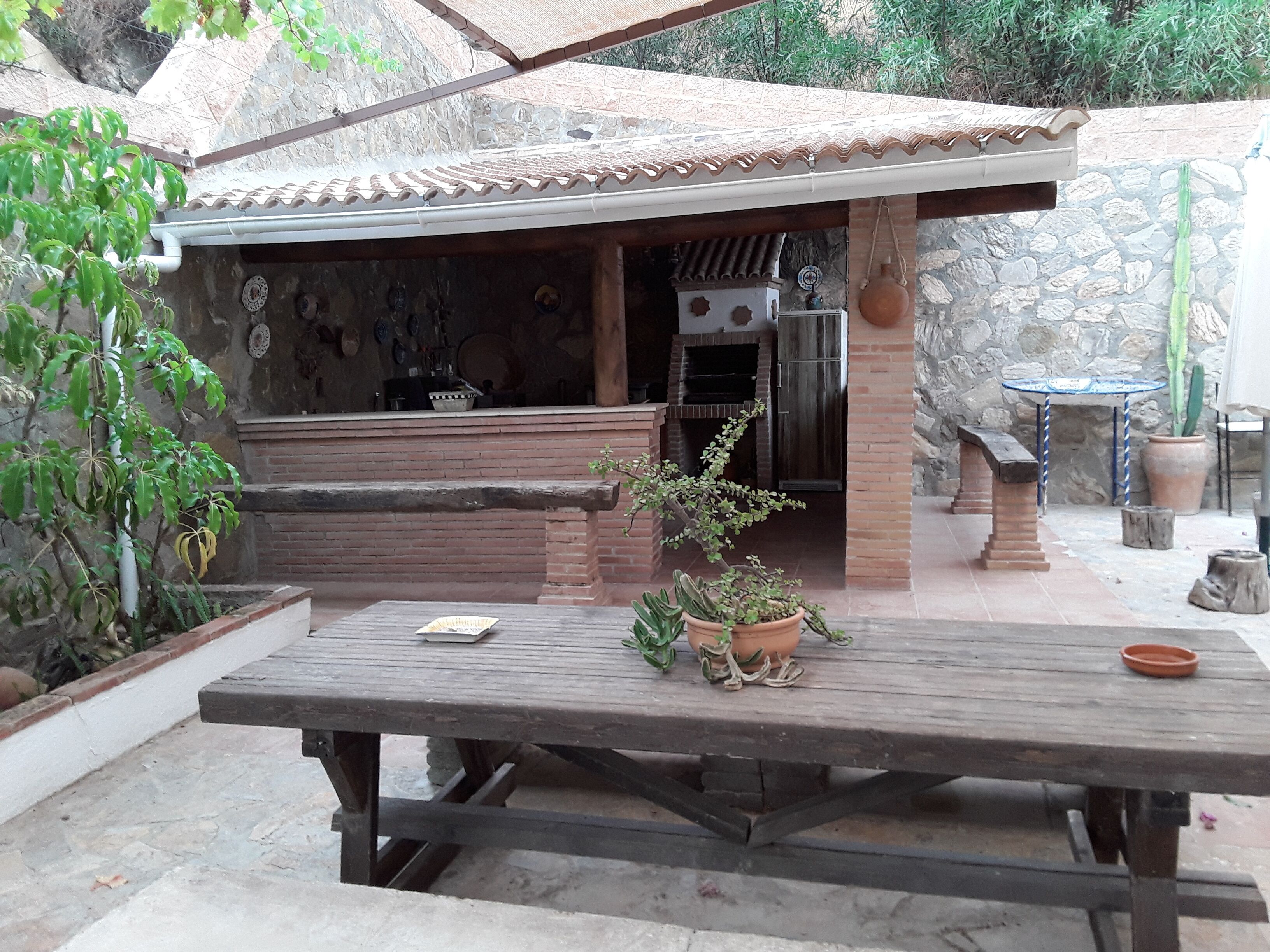 Une terrasse extérieure avec une table en bois, un espace barbecue en briques et des plantes en pot.