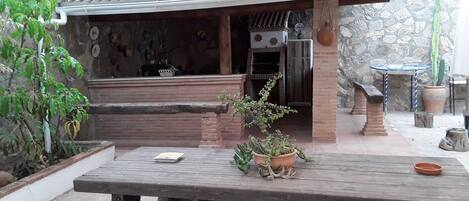 Une terrasse extérieure avec une table en bois, un espace barbecue en briques et des plantes en pot.