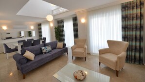 Un salon moderne avec un canapé gris, des fauteuils beige et une table basse en verre. On aperçoit à l’arrière-plan un coin repas avec une table et des chaises blanches.