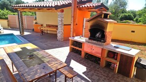 An outdoor barbecue area with a brick oven, a pool, and a dining setup. - CASA BOZZERO, elegant and modern rustic with 2 apartments (Krnica)