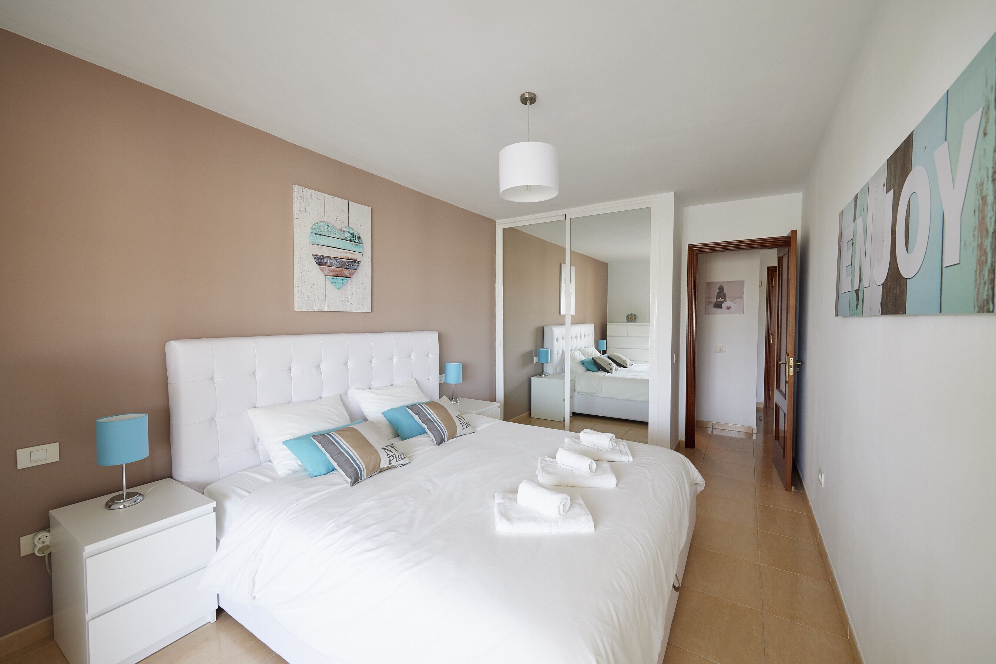 A neatly arranged bedroom with a large bed, a nightstand, a mirror, and a wall with a 'Enjoy' sign.