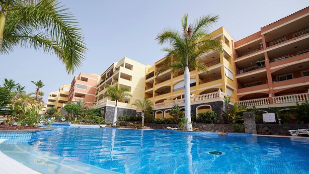 A swimming pool with a tiled deck, surrounded by multi-story buildings with balconies and palm trees. - Spacious and cozy apartment  (Palm-Mar)