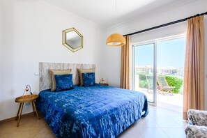 A bedroom with a large bed, a round side table, a hanging light fixture, and a view of the outside.