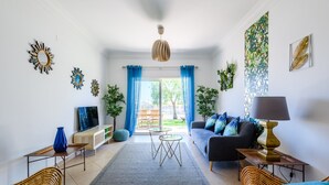 Una sala de estar con un sofá, una mesa de centro y un televisor. Hay cortinas, una planta y una obra de arte en la pared.