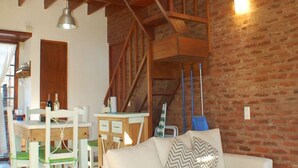 Amplia sala de estar con techo de madera alto, un sofá blanco con cojines estampados, una mesa de centro de madera y un comedor con mesa y sillas de madera.