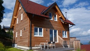 A wooden house with a red roof, a covered patio with outdoor furniture, and a view of the surrounding landscape. - Biological holiday above the clouds of Carinthia in every season (Hochrindl-Kegel)