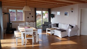 A spacious living area with a wooden ceiling, a white sofa, a dining table with chairs, and a large window offering a view of a red-roofed building. - Biological holiday above the clouds of Carinthia in every season (Hochrindl-Kegel)