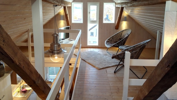 Un dormitorio acogedor en el ático con vigas de madera, un escritorio con lámpara, dos sillones de mimbre y vistas al exterior a través de amplias ventanas.