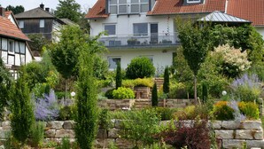 Una casa de dos pisos con balcón, rodeada por un jardín bien cuidado con diversas plantas y un muro de piedra.