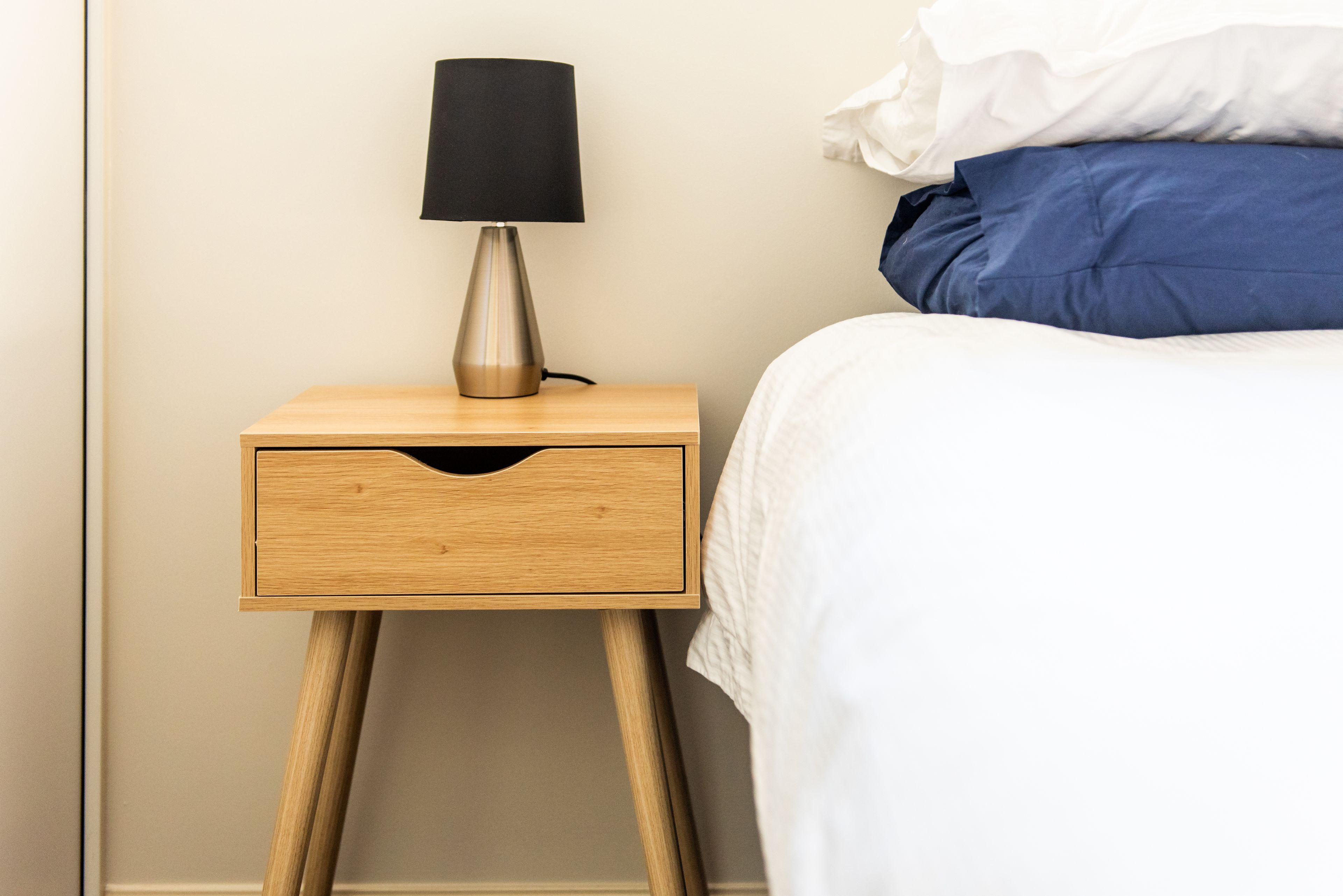 A wooden bedside table with a lamp, a drawer, and a bed with white and blue bedding.