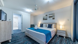 Habitación de hotel con cama, escritorio, silla, televisor y baño visible a través de una puerta abierta.