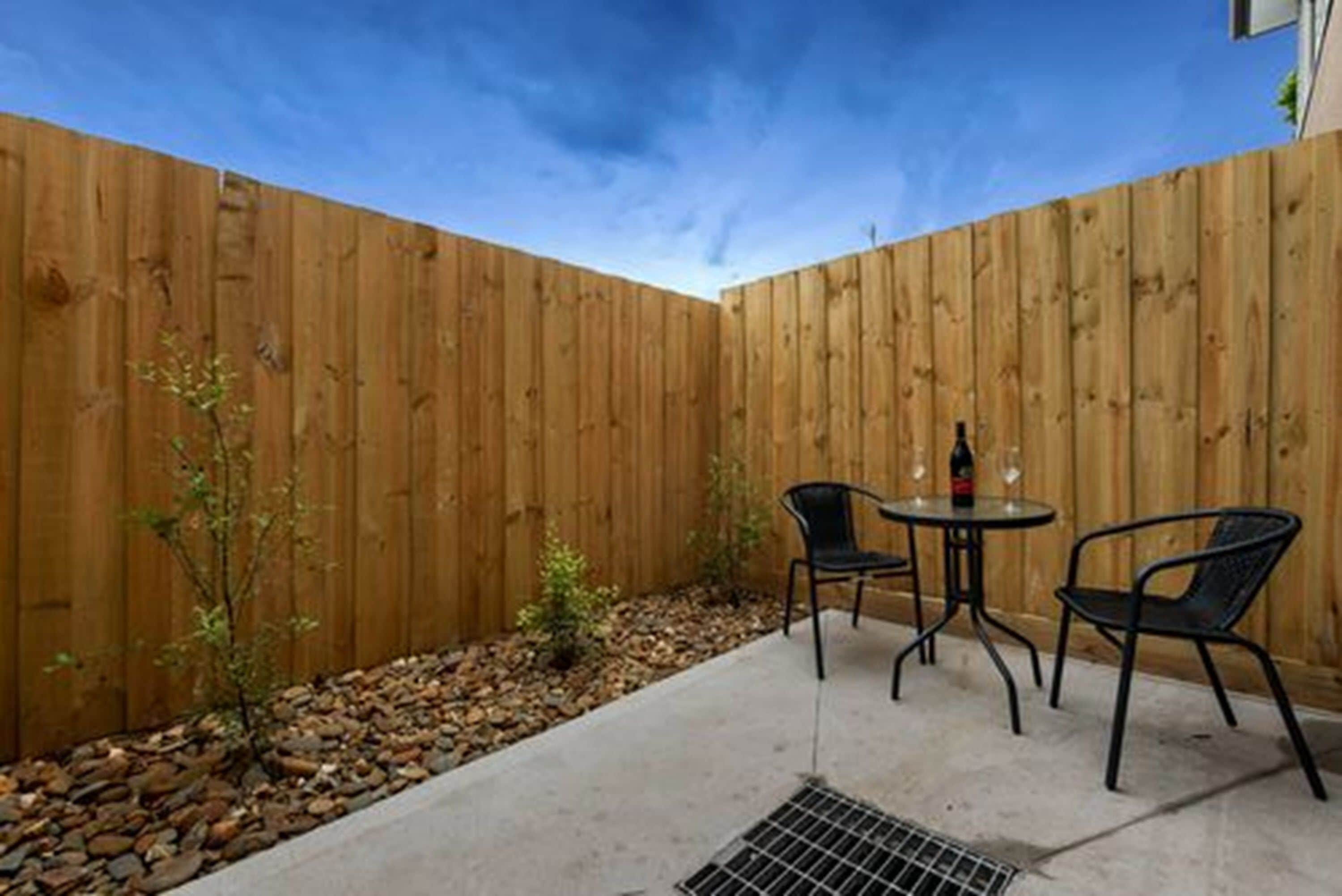 A patio area with a wooden fence, two chairs, a small table, and a bottle of wine.