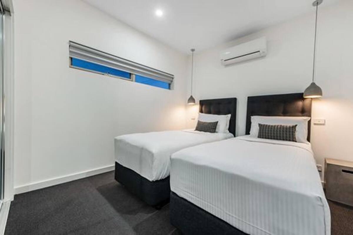 A hotel room with two single beds, a window with blinds, an air conditioning unit, and a pendant light.