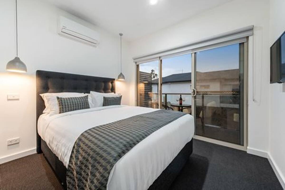 A modern hotel room with a large bed, a sliding glass door to the balcony, and a flat-screen TV mounted on the wall.
