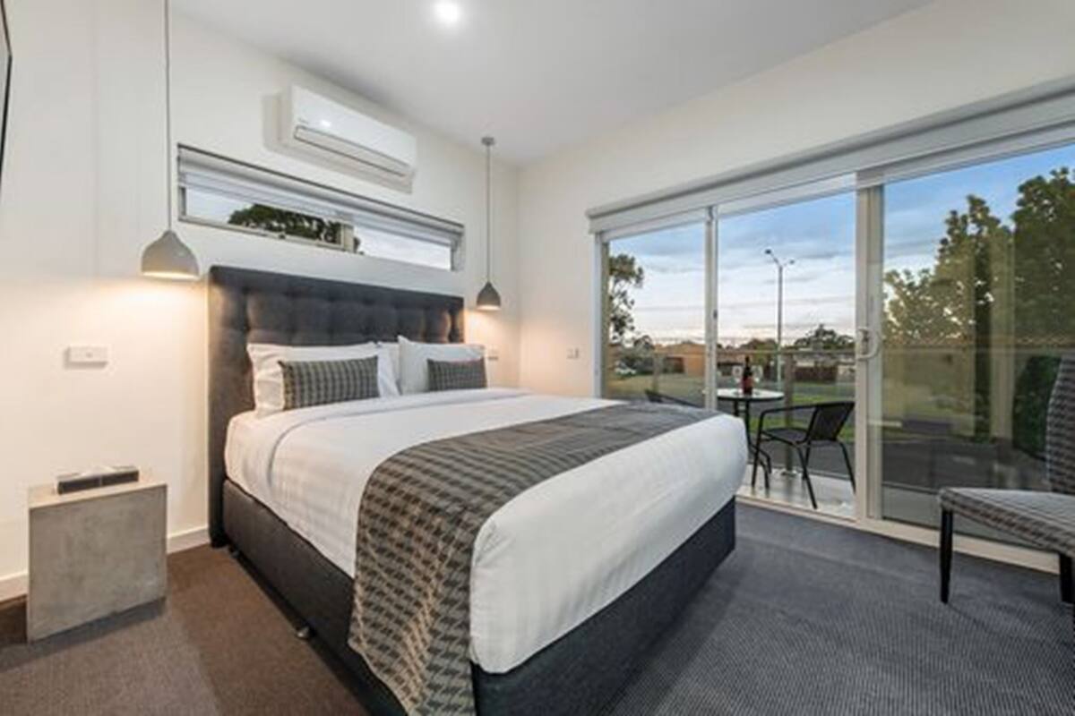 A modern bedroom with a large bed, a small bedside table, a bench, and a sliding glass door leading to an outdoor area.