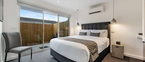 A modern bedroom with a large bed, a chair, and a sliding glass door leading to a backyard.