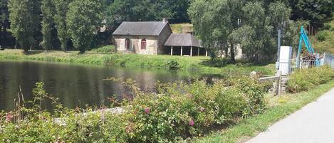 A house by a lake with a stone exterior, surrounded by lush greenery and a clear sky.