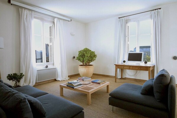 A living room with two sofas, a coffee table with books, a potted plant, a desk with a computer, and large windows with white curtains. - Air conditioned duplex in a 19th century mansion centrally located with parking (Tours)