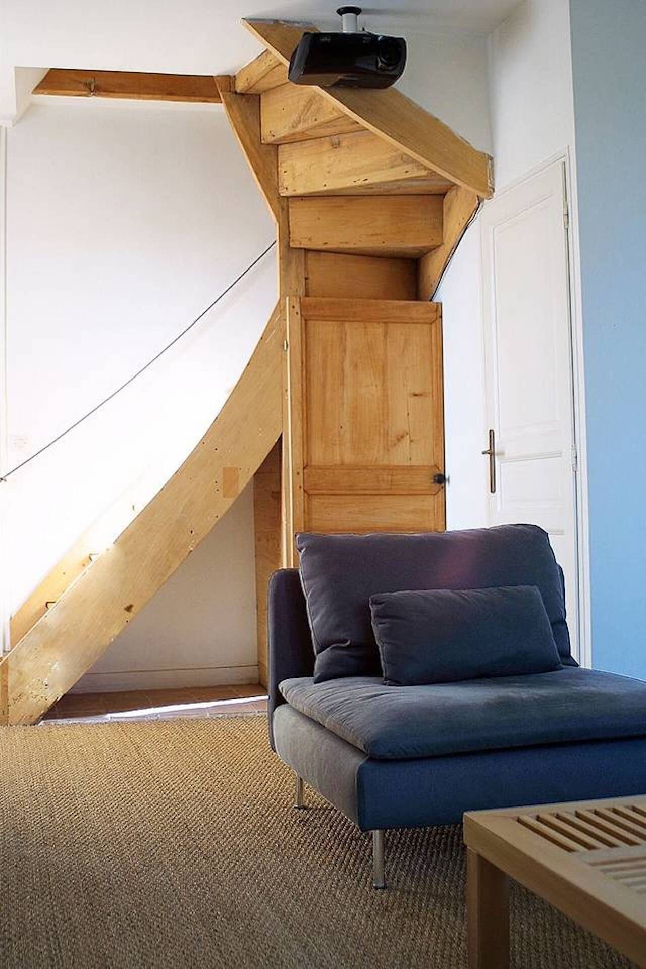 A room with a wooden staircase leading to an upper level, a blue sofa, and a wooden table.