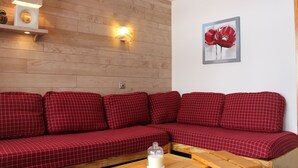 Un sofá rojo con estampado de cuadros, una mesa de madera al frente, una lámpara de pared y un cuadro con flores rojas.