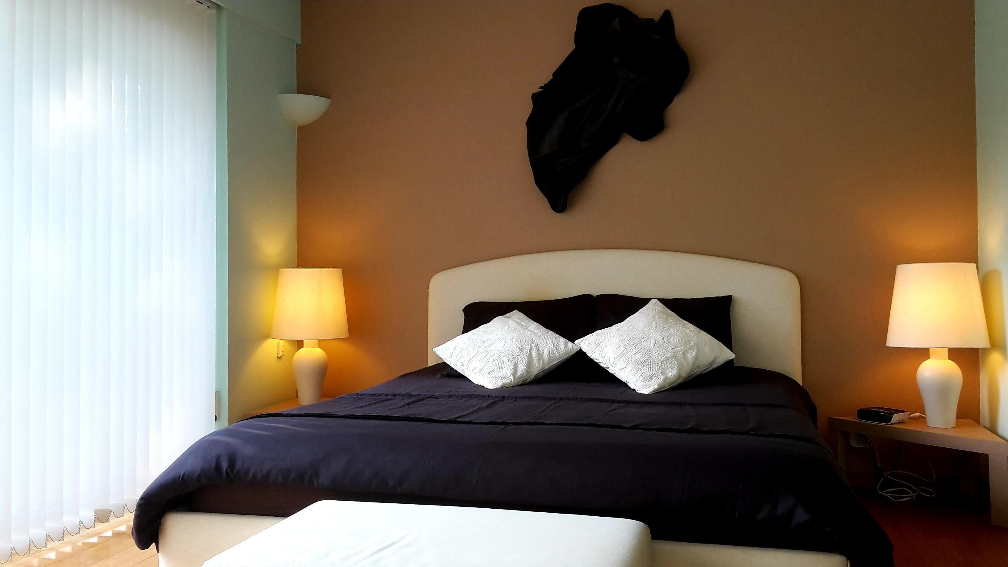A hotel room with a bed, two bedside lamps, a black coat hanging on the wall, and a window with blinds.