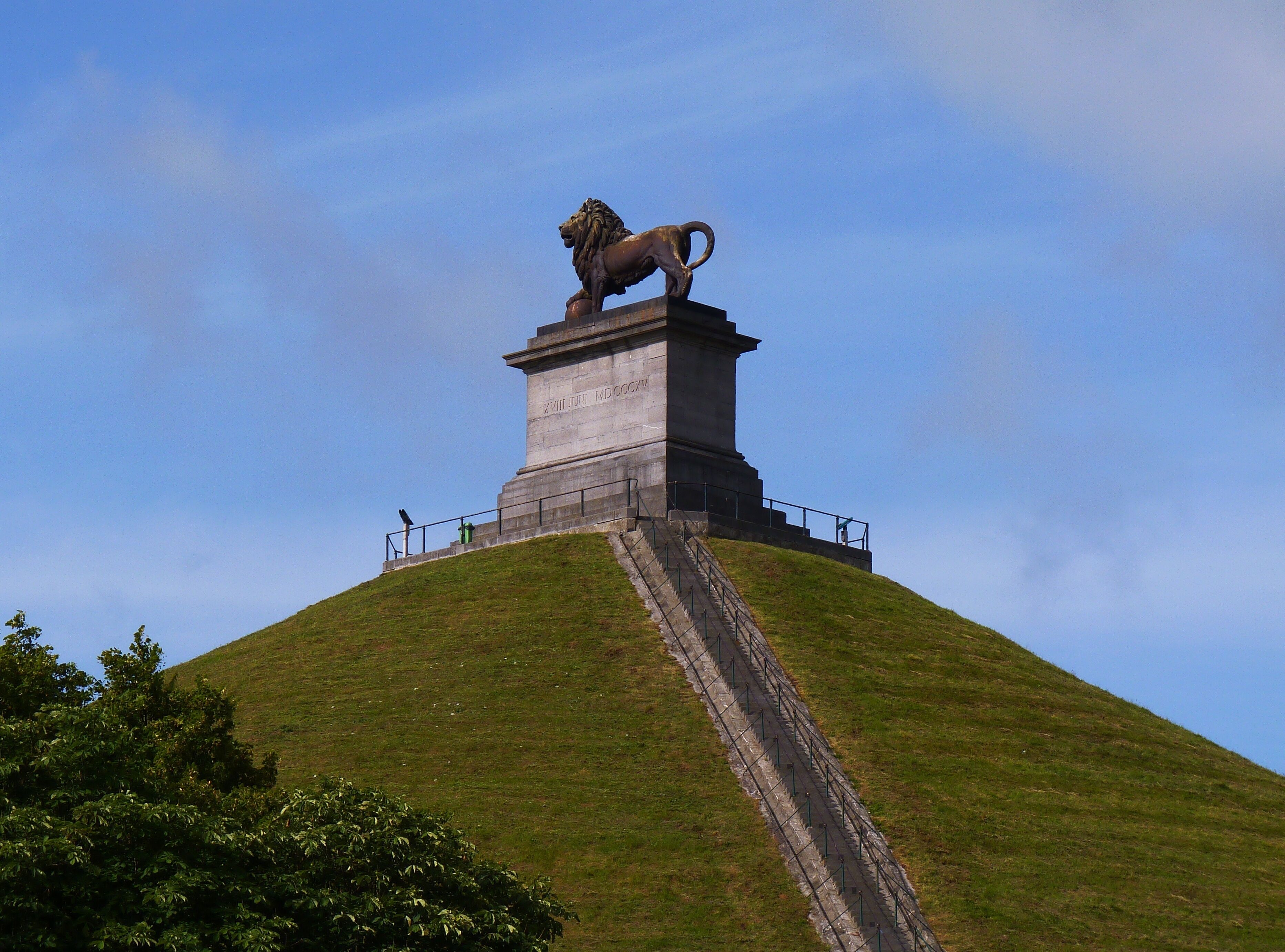 Een monument met een leeuwenbeeld bovenop, omgeven door een grasheuvel en een heldere lucht.