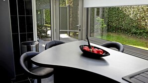Ein moderner Essbereich mit einem gewölbten Tisch und schwarzen Stühlen, eine Schale mit roten Äpfeln auf dem Tisch und Blick auf einen Garten durch große Fenster.