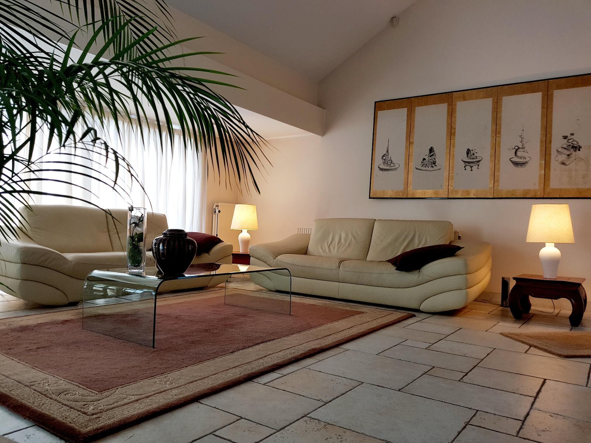 Een woonkamer met een beige bank, een glazen salontafel, een rood kleed, een potplant en een muur met ingekaderde kunstwerken.