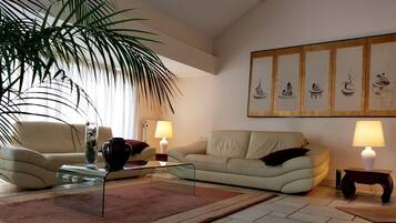 Een woonkamer met een beige bank, een glazen salontafel, een rood kleed, een potplant en een muur met ingekaderde kunstwerken.