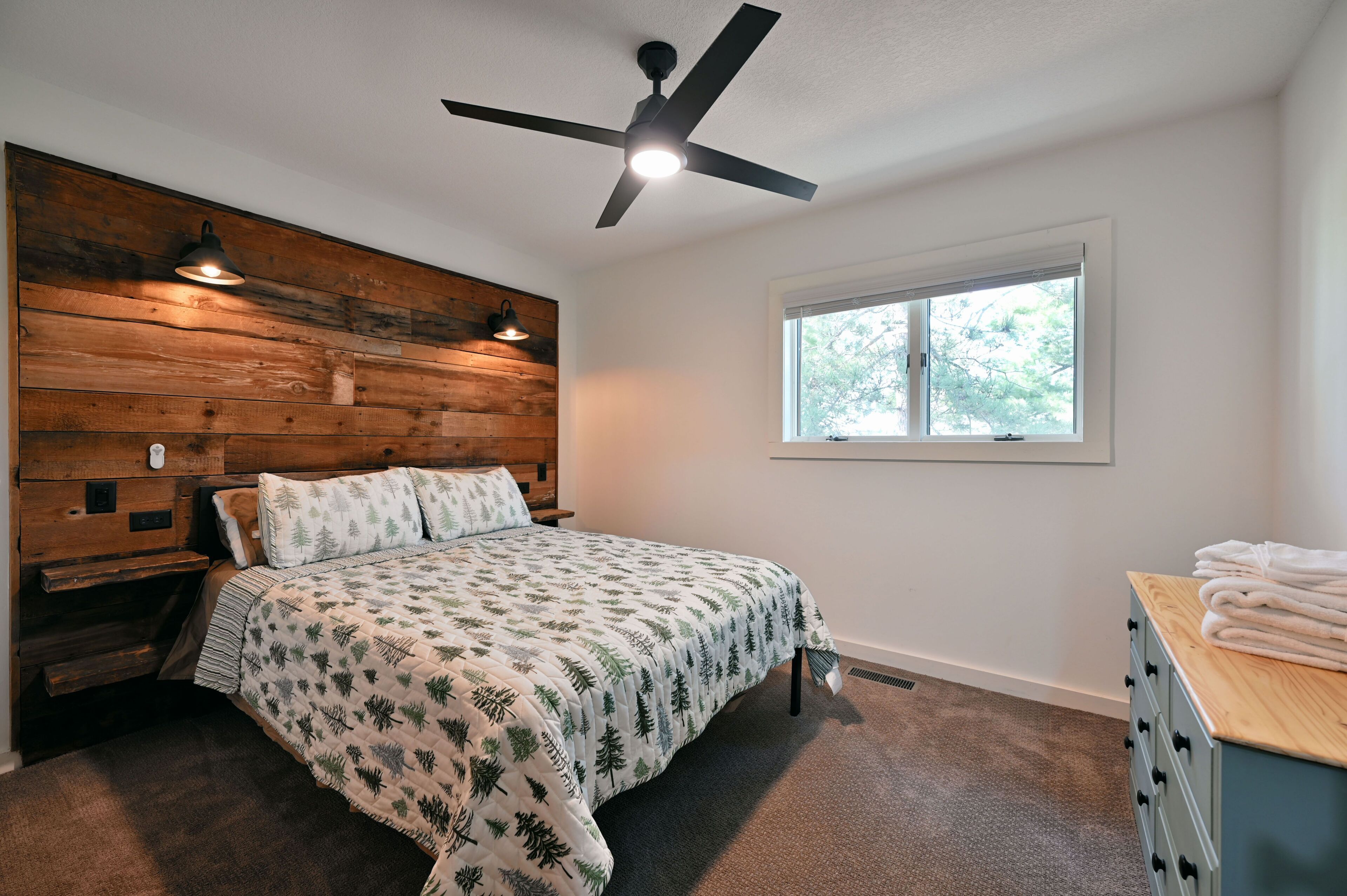 A bedroom with a wooden headboard, a bed with a leaf-patterned bedspread, a ceiling fan, a window with a view of trees, and a dresser with towels.