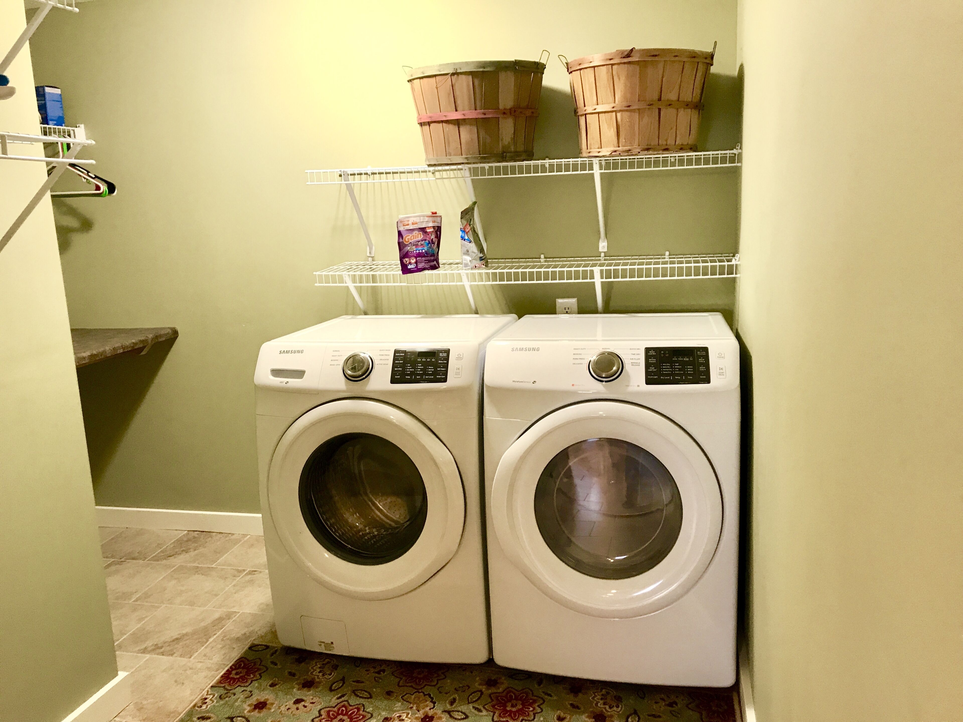 Un espace buanderie comprenant deux machines à laver Samsung blanches, une étagère avec des paniers et une boîte de lessive.