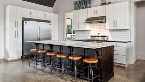 A modern kitchen with white cabinetry, a large island with stools, and a stainless steel refrigerator. - Modern Lake House on Smith Lake, AL close to Birmingham and Huntsville (Bremen)