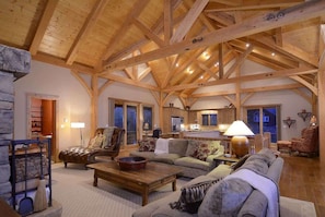 A spacious living area with a high wooden ceiling, a fireplace, a sofa, a coffee table, and a kitchen in the background.