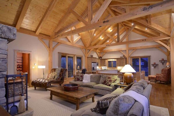 A spacious living area with a high wooden ceiling, a fireplace, a sofa, a coffee table, and a kitchen in the background.
