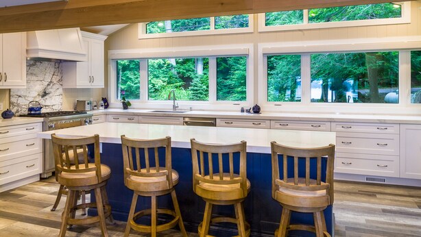 A modern kitchen with a large island, white cabinetry, and a view of the forest through large windows. - Lake Rosseau - Rare, Flat double lot with breathtaking South West views. (Minett)