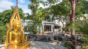 Een gouden Boeddhabeeld in een meditatieve houding, omringd door een stenen terras en palmbomen, met een zwembad en ligstoelen op de achtergrond.