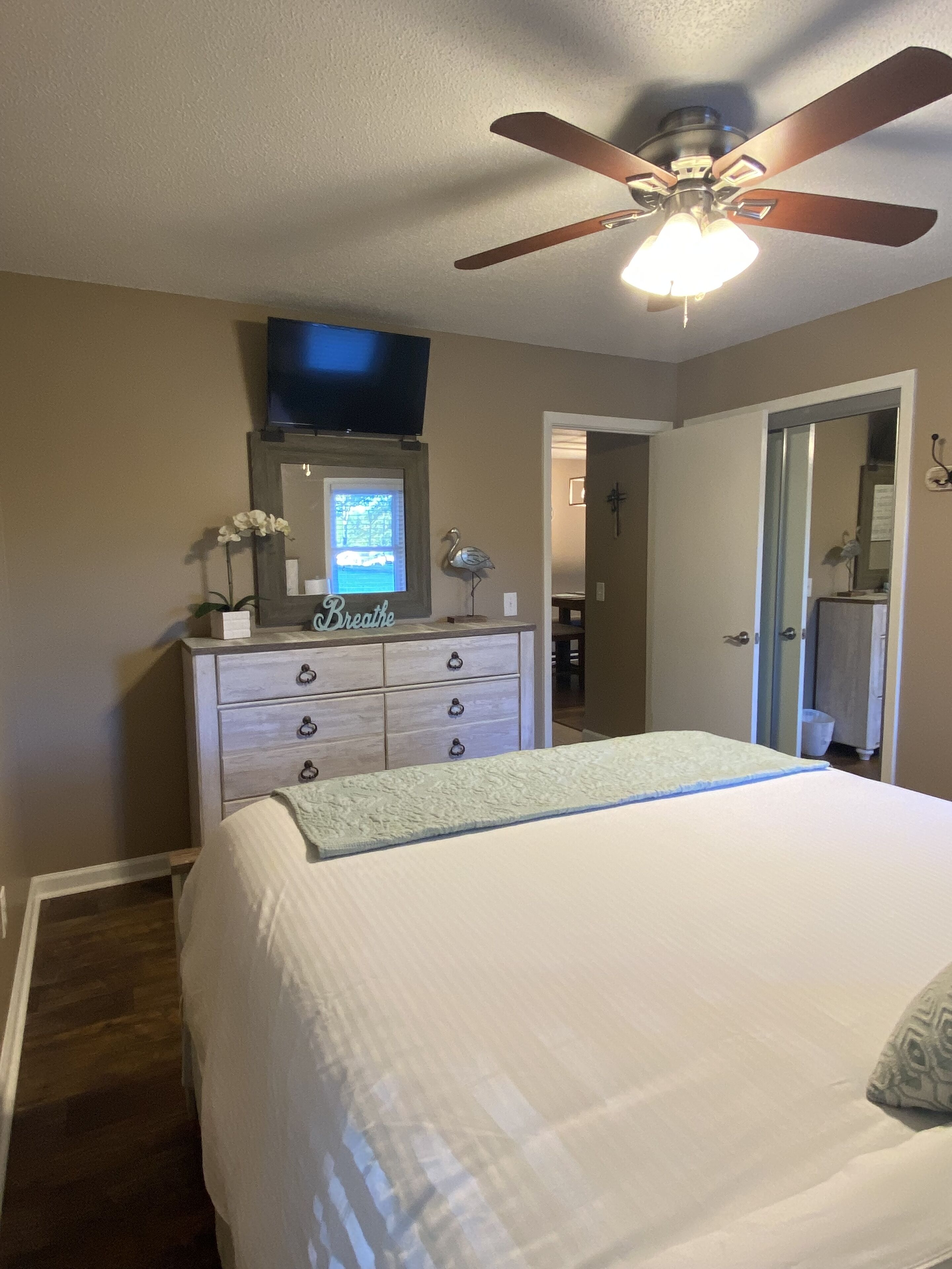 A bedroom with a bed, dresser, ceiling fan, and a TV mounted on the wall.
